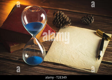 Hourglass and old books with old paper and pen on wooden tables in lighting of the lantern. Stock Photo