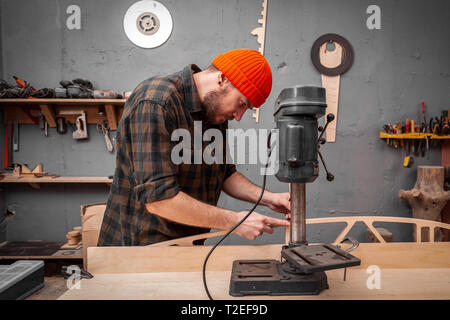 A man carpenter in a hat and a shirt  is carving a wooden board on a large drilling machine in a workshop side view, in the background a lot of tools Stock Photo