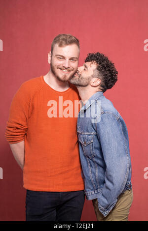 two men, gay couple posing together, smiling and happy. one is looking directly to camera. other is kissing him in the cheek. Stock Photo