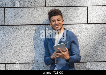Young mulatto man standing isolated on concrete wall holding digital tablet browsing internet looking camera smiling joyful Stock Photo