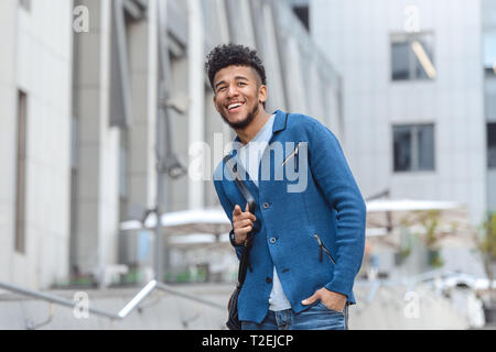Young mulatto man carrying backpack standing on city street pointing to camera laughing playful Stock Photo