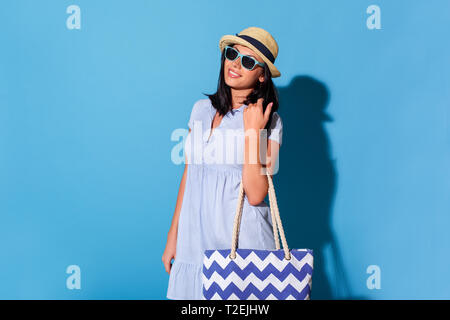 Young woman wearing dress sunglasses and hat standing isolated on blue wall carrying bag looking camera smiling cheerful Stock Photo