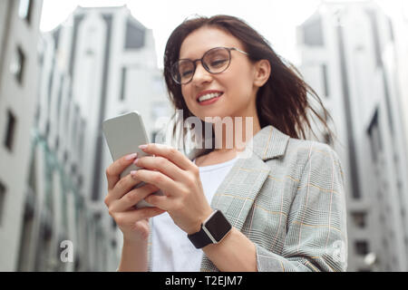 Young businesswoman wearing eyeglasses standing on the city street using application on smartphone smiling cheerful close-up blurred Stock Photo