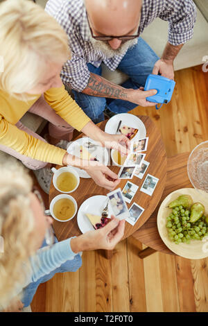 Directly above view of mature friends sitting on sofa and talking while watching images made at party, they drinking tea and eating cake Stock Photo