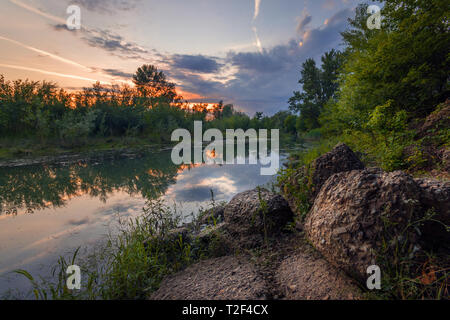 Vibrant sunset scene near a pond surrounded by trees with rocks in the foreground Stock Photo