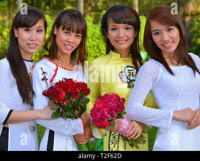 Four dressed-up Vietnamese girls wear traditional silk dresses (áo dài) and pose with two bunches of roses for the camera.