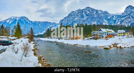 The winter landscape of Obertraun village with huge mountains of Dachstein massif, old coniferous forest, traditional houses and Traun river, Salzkamm Stock Photo