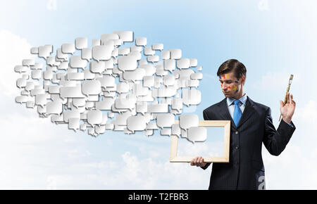 Portrait of young and successful businessman in black suit holding paintbrush in hands an looking at picture frame while standing against blue cloudy  Stock Photo