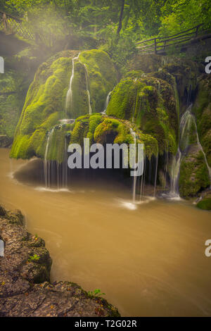 Beautiful waterfall set in a scenic landscape with a big cliff covered in moss falling into a river with a bridge behind and some rocks in the foregro