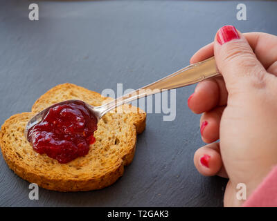 A woman with painted nails smearing red raspberry jam with a spoon on a slice of toast Stock Photo