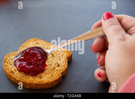 A woman with painted nails smearing red raspberry jam with a spoon on a slice of toast Stock Photo