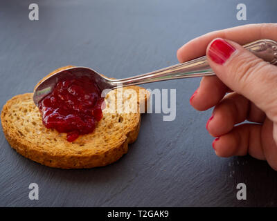 A woman with painted nails smearing red raspberry jam with a spoon on a slice of toast Stock Photo