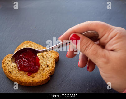 A woman with painted nails smearing red raspberry jam with a spoon on a slice of toast Stock Photo