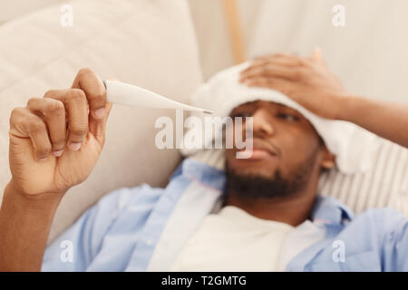Fever, seasonal flu. Sick african-american man lying on sofa and checking his temperature with thermometer, free space Stock Photo