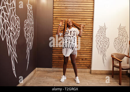 Beautiful african woman in stylish casual clothes posing against angel wings wall. She moving her hair dreadlocks. Stock Photo