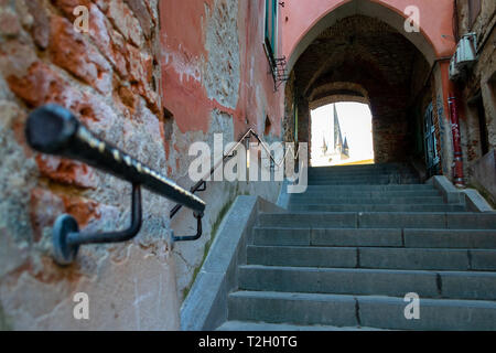 Old narrow street with shriveled brick walls, cement stairs and a black metal handrail pointing up towards an archway with a a few church towers in th Stock Photo
