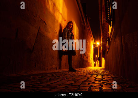 Moody portrait of a young woman on a narrow urban passage, at night, next to open window shutters, in Sibiu (Hermannstadt), with scarce street lights  Stock Photo