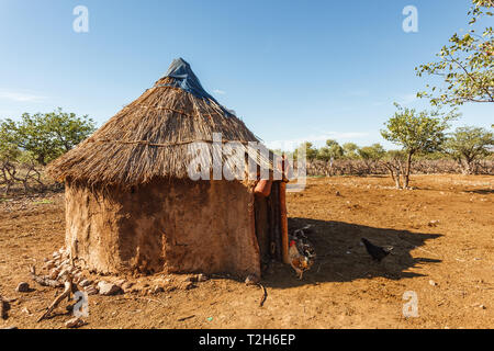 Closeup of round mud hut with straw roof  in Himba village and chickens by the door in Namibia, Africa Stock Photo