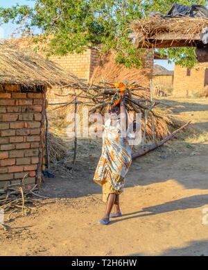 Malawian woman carries firewood on her head with her baby on her back outside her kitchen in a village Stock Photo