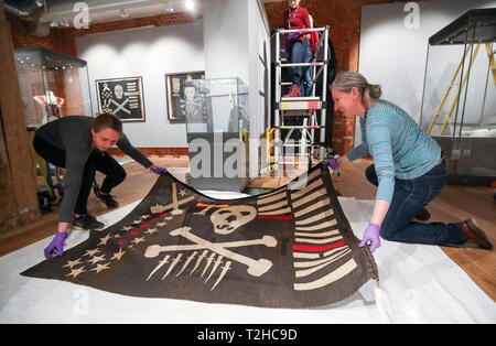 Victoria Ingles (right), senior curator at the National Museum of the Royal Navy, helps un-roll a Jolly Roger once flown by HMS Safari during World War 2, during the preview of Jolly Roger: A Symbol of Terror and Pride exhibition at the National Museum of the Royal Navy at Portsmouth Historic Dockyard. Stock Photo