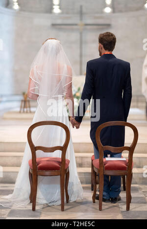 Religious wedding in a church: couple of newlyweds standing, holding hands, facing a cross in the background Stock Photo