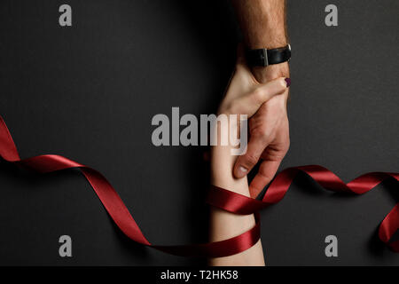 cropped view of man and woman in red satin ribbon holding hands on black background Stock Photo