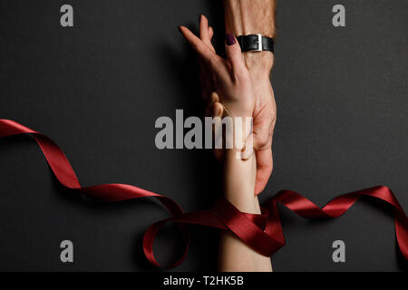 cropped view of man holding female hand in red satin ribbon on black background Stock Photo