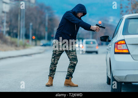 Gangster with a gun trying to rob a guy which is sitting in his car. Stock Photo