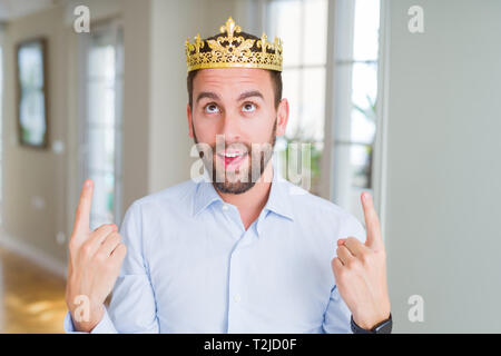 Handsome business man wearing golden crown as a king or prince amazed and surprised looking up and pointing with fingers and raised arms. Stock Photo