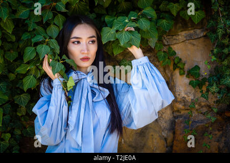 Portrait of young stylish asian woman in green leaves on stone wall background Stock Photo