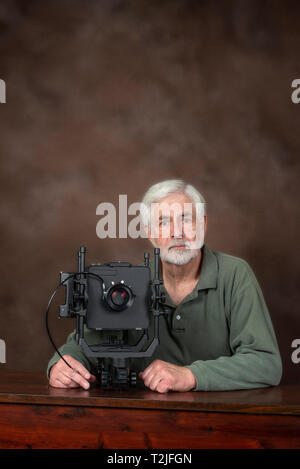 Vertical shot of an elderly photographer sitting behind a professional view camera.  Brown background with lots of copy space above his head. Stock Photo