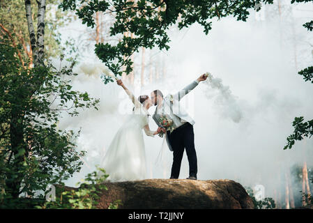 The bride and groom stand on a cliff and hold smoke bombs in their hands in white. Stock Photo