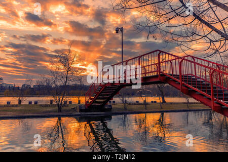 Sunset seen from a park in Bucharest with sun rays coming out from the clouds and a red metal bridge in the foreground shot at the beginning of the sp Stock Photo