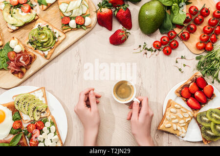 top view of woman holding cup of coffee at table with fresh ingredients, greenery and toasts with vegetables and prosciutto Stock Photo
