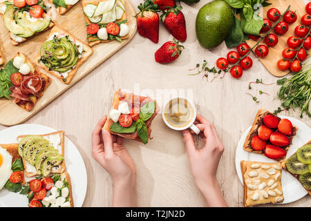 top view of woman holding cup of coffee at table with ingredients, greenery and toasts with vegetables and prosciutto Stock Photo