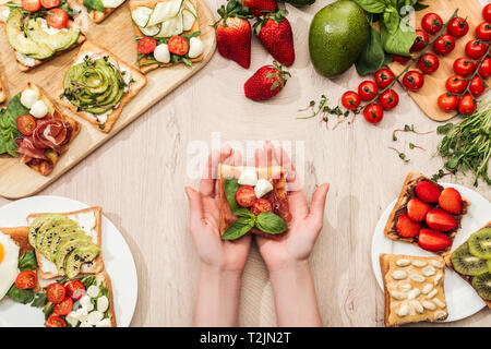 top view of woman holding toast with cherry tomatoes and prosciutto over wooden table with greenery and ingredients Stock Photo