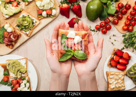 selective focus of woman holding toast with cherry tomatoes and prosciutto over wooden table with greenery and ingredients Stock Photo