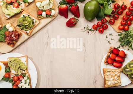 top view of toasts with vegetables, fruits and prosciutto with greenery and ingredients on wooden table Stock Photo