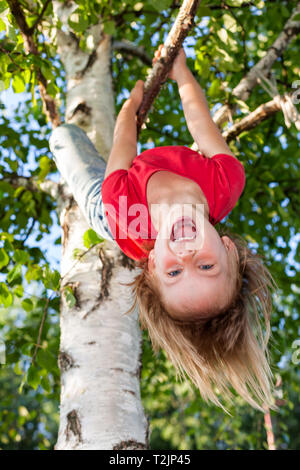 Portrait of happy elementary age girl wearing red tee shirt hanging upside down from a tree branch - summer fun concept Stock Photo