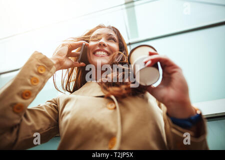 Beautiful young woman with a disposable coffee cup, drinking coffee and talking on a smartphone against urban city background. Stock Photo