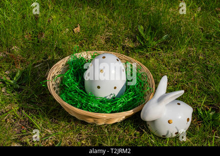 An Easter bunny and an Easter egg made of white pottery with golden polka dots with a basket on a meadow. Stock Photo