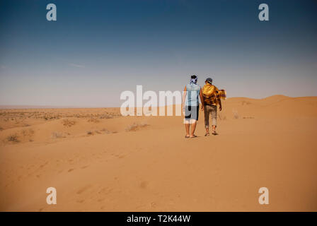 There are two men walking on hot desert sands together toward the blue sky under natural burning sun Stock Photo