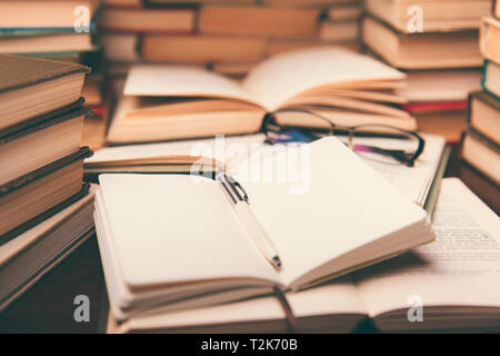 Education learning concept with open book and notebook on the table. Stack piles of books on reading desk and glasses in bookshelves background Stock Photo