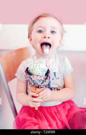 Cute funny Caucasian blonde babyl girl child with blue eyes eating licking ice cream in large waffle cone with sprinkles and showing tongue. Happy chi