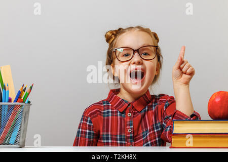 Funny little preschool girl in glasses sits at the table with books and points finger up. The concept of education. Gray background. Stock Photo