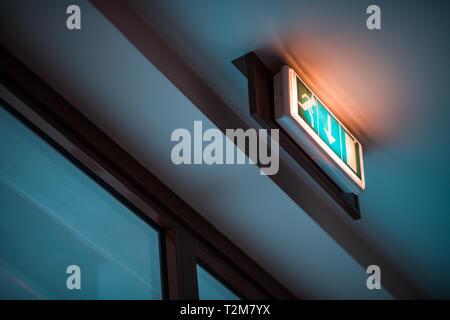 Signal and green luminous emergency exit sign hanging from the ceiling above a door inside a public building Stock Photo