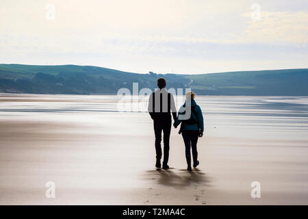 A couple holding hands walking together on a quiet sandy beach at low tide. Woolacombe, North Devon, England, UK, Britain Stock Photo