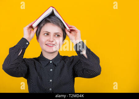 Happy young woman dreamer looks aside with positive expression, keeps open book over head, wears black shirt, demands holidays, isolated over yellow w Stock Photo