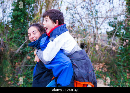 oung boy piggyback on his sister looking at camera and smiling in nature Stock Photo