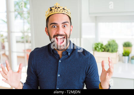 Handsome hispanic man wearing golden crown over head as the king celebrating crazy and amazed for success with arms raised and open eyes screaming exc Stock Photo